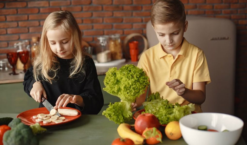 Focused little girl and boy sitting at desk with assorted vegetables and fruits while cooking in loft kitchen with brick wall