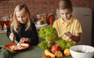 Focused little girl and boy sitting at desk with assorted vegetables and fruits while cooking in loft kitchen with brick wall