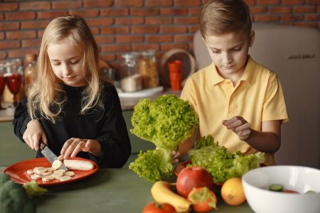 Focused little girl and boy sitting at desk with assorted vegetables and fruits while cooking in loft kitchen with brick wall